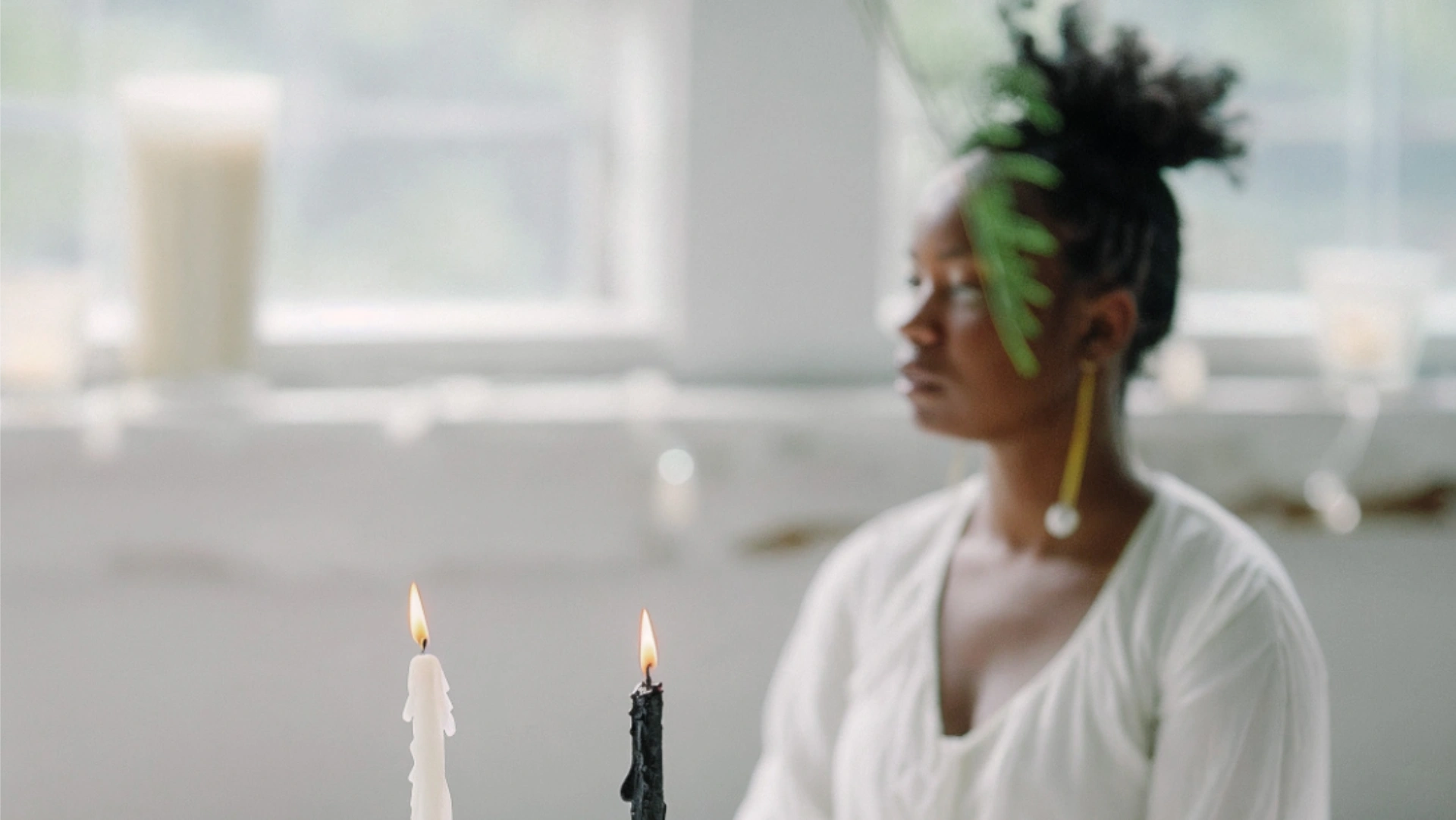 Soft-focus image of a woman in white sitting peacefully in a sunlit room, with a white and black candle burning in the foreground.