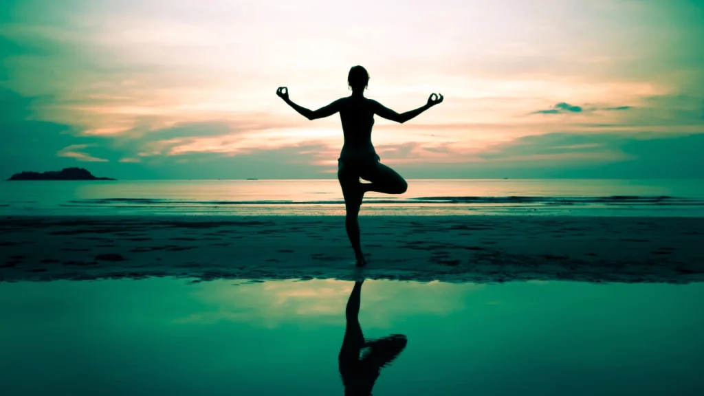 Silhouette of a woman practicing yoga on the beach at sunrise, symbolizing balance, inner peace, and the whole-person approach of holistic health care.