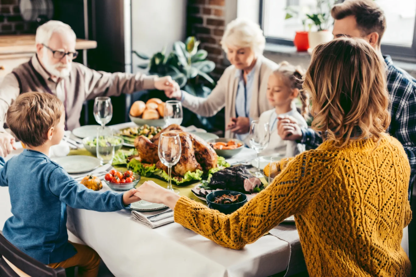 A multigenerational family sitting around a holiday table, holding hands before a meal that includes turkey, vegetables, and bread rolls, symbolizing gratitude and togetherness during the holidays.