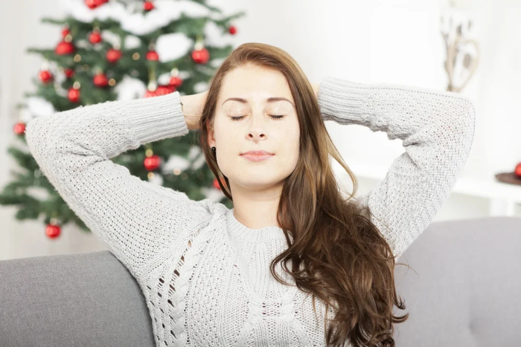 Woman sitting on a couch with her eyes closed and hands behind her head, taking a calming breath with a decorated Christmas tree in the background.