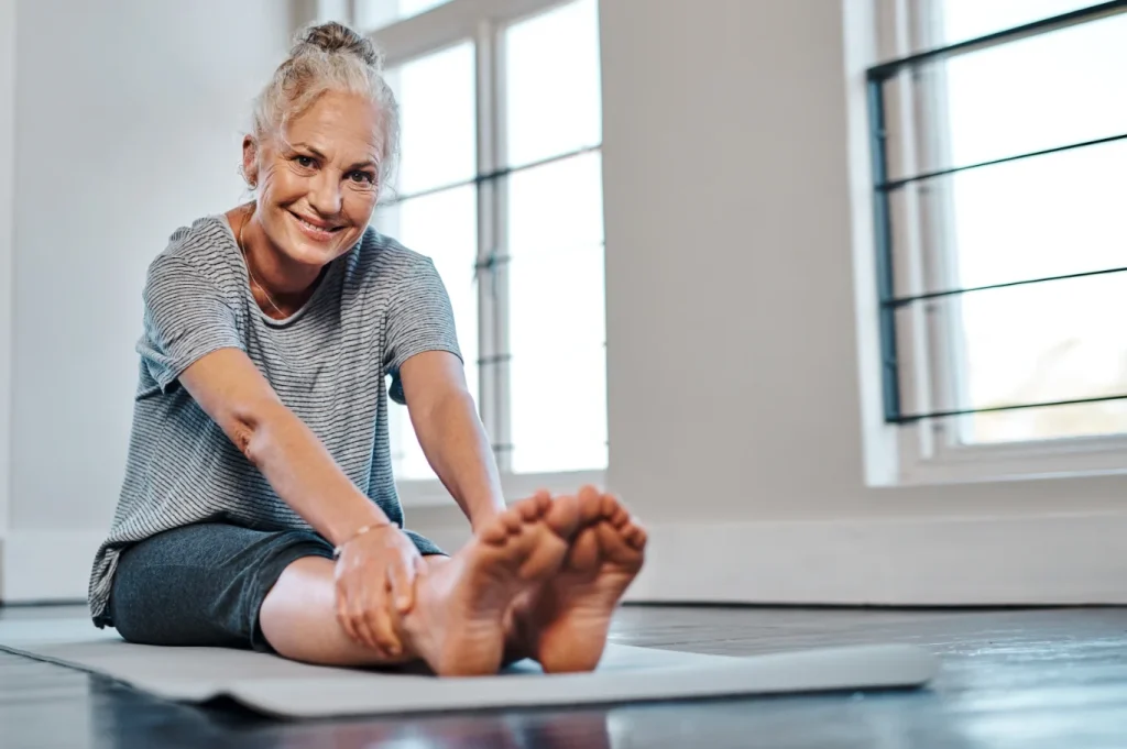 Woman seated on a yoga mat indoors, stretching her legs during a gentle wellness or movement practice.