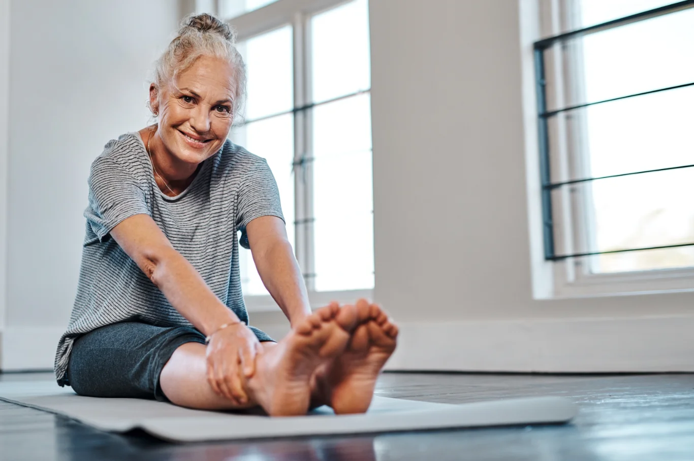 Woman seated on a yoga mat indoors, stretching her legs during a gentle wellness or movement practice.